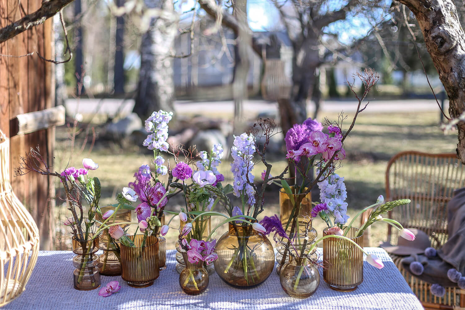 Puutarhapöydälle on tehty kukka-asetelma johon on koottu erilaisia ruskean sävyisiä maljakoita ja niissä on liloja kukkia.